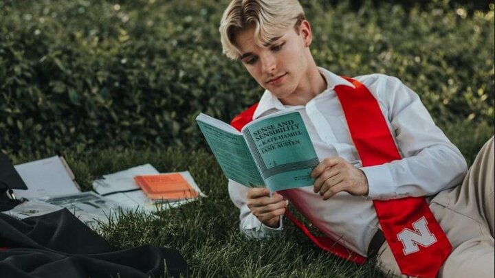 Student, Gage Boardman, wearing red graduation sash, laying down on grass reading a script book.