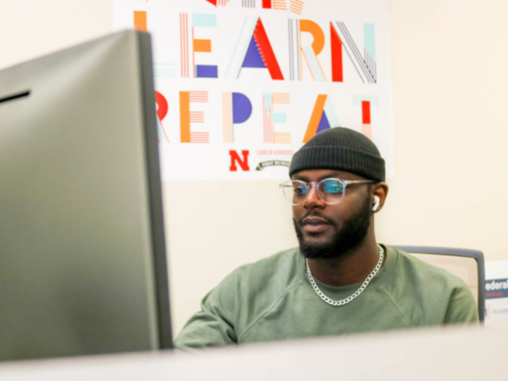 student wearing glasses and cap in front of computer screen